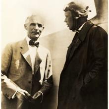 Charles Lindbergh and Henry Ford, both of whom are white men, stand together. Lindbergh is wearing aviator gear.