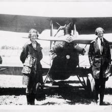 Neta Snook, a female flight instructor, stands with her at-the-time female pilot student, Amelia Earhart, in front of a biplane with one engine.