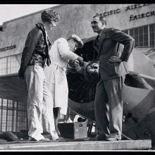 Paul Mantz, a man, stands to the right of Amelia Earhart, a white woman, as they speak to each other.