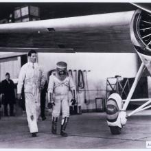 Wiley Post walks under his monoplane, wearing a pressure suit he planned to test while flying his aircraft. The pressure suit features a cylinder-shaped helmet and dark-colored boots.