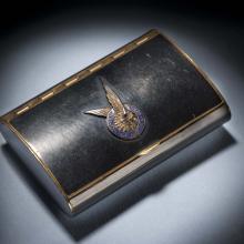 Black makeup case with closed lid. On the center of the lid, there is a silver insignia of a pair of two wings inside a blue seal.