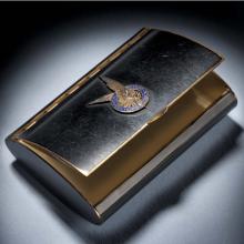 Black makeup case with open lid. On the center of the lid, there is a silver insignia of a pair of two wings inside a blue seal.