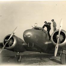 Amelia Earhart and Fred Noonan, a white woman and white male respectively, stand on top of a monoplane with twin engines. Noonan is climbing into the monoplane.