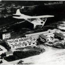 A monoplane flies over a set of buildings which are labeled as belonging to the "Taylor Aircraft Co." on the roof of a building on the left of the other buildings. Below the roof stating the company's name is the location of "Bradford, PA" on a lower roof.