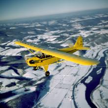 A yellow monoplane with one engine flies above land.