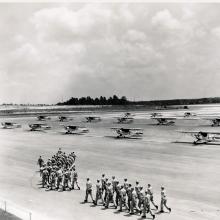 Multiple lines of Piper Cubs, monoplanes with one engine, are seen on a U.S. Navy training site. Groups of men are walking in formation in front of the planes.