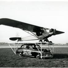 A Piper Cub, a monoplane with one engine, is placed on top of a car prior to performing a stunt.