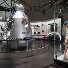 Side view of an exhibit about a Red Bull-sponsored free falling mission from the edge of space. A high-altitude pressure suit is seen in front of the silver-colored opened capsule used to freefall.