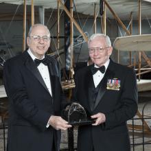 Two white men stand together, holding a trophy award at a Museum awards event.