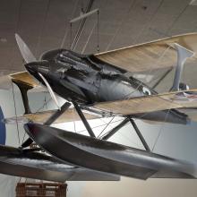 Diagonal and frontal view of black biplane with single engine and pontoon floats hanging in the Museum.