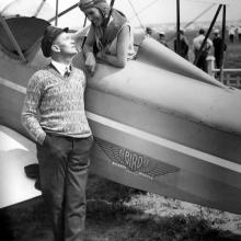 A woman in a pilot's cap leans out of a plane. A man standing on the ground stares up at her. The photo is in black and white.