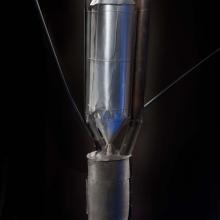 Close-up view of motor on a primitive rocket. A conical nose tops a metal cylinder-shaped motor attached to a cylinder-shaped fuel tank by a metal conical-shaped drain.