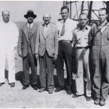 Six white men, including Harry Guggenheim, Robert Goddard, and Charles Lindbergh, stand in front of a tower in a desert.