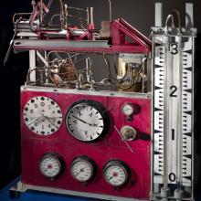 Large table stand with multiple clock-like meters and a large pressure meter. Above the meters are a two-story set of tubes.