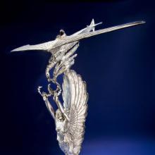 Silver-colored trophy with topper of a winged angel holding olive wreaths which are picked up by a plane.