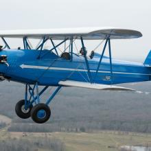 Side view of blue biplane with white wings, one engine, and fixed landing gear. Biplane is in flight. Registration number "N980V" painted in white paint on rudder.