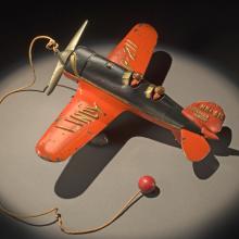 Top view of orange and black monoplane toy with a ball and string attached to the center of the propellors on the front.