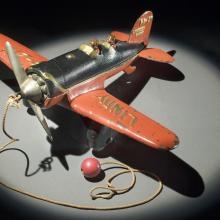 Top view of orange and black monoplane toy with a ball and string attached to the center of the propellors on the front.