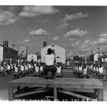 Partial view of some of the buildings and a clearing at the Tuskegee Army Air Field. A person runs physical training for cadets in the foreground.