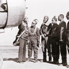 Five pilots who recently received their new status as Tuskegee pilots stand with another person next to a trainer aircraft.