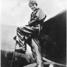 Amelia Earhart, a white woman, climbs into the cockpit of a plane.