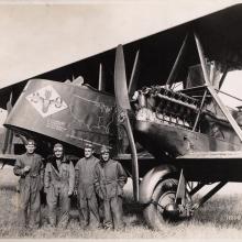 A group of white males representing the U.S. Army Air Service stand next to the nose of a biplane.