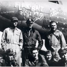 A group of seven members of a bomber combat plane used in World War II named "Flak-Bait" pose formally in front of the aircraft following a milestone mission.