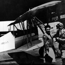 Chauncey Spencer, an African-American male pilot, stands next to a biplane with another person.