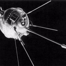 View of metal orbiting sphere-like base of an object with multiple antenna probes attached to bottom of base object. A square attachement featuring smaller square panels is attached above the antennae probes.