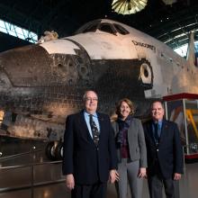 Three people, two white men and one white woman, stand formally in front of the Space Shuttle Discovery, a white spacecraft.