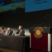 Five people present a lecture at the Museum. One is standing next to a podium whereas four other people are sitting at a table.