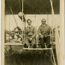 Harry Bingham Brown, a white male pilot, sits in his Wright biplane on the left of another male. Brown is smoking a cigar.