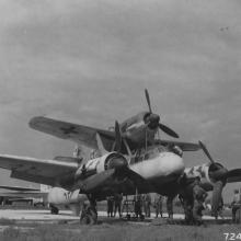 Men stand around a aircraft with two engines which is holding a smaller monoplane with one engine on top of the fuselage.