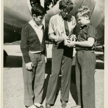 Amelia Earhart, a white woman, stands in between two white people in front of an aircraft. She is signing an autograph for the person on her left (our right).