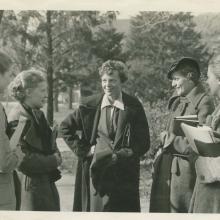 Amelia Earhart, a white female pilot, stands in the center of a group of five people. She is smiling toward the people on the left.