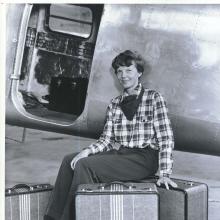 Amelia Earhart, a white female pilot, poses in front of an aircraft with three pieces of luggage that she designed.