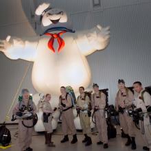 People dressed up in Ghostbusters costumes stand together in front of a Ghostbusters ghost float inside the Museum.