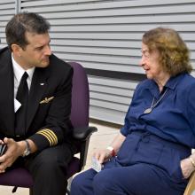 Jerrie Mock, a white female pilot, sits next to and speaks to a male pilot at a Museum event.