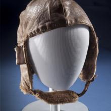 Brown leather aviation helmet with brown fur-lined chin strap. Styled on mannequin.