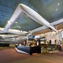 Two docents stand at the museum's welcome desk. A white, twin-boom aircraft hangs above the desk.