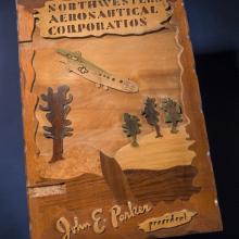 Scrapbook cover featuring a depiction of an aircraft in a downwards position moving toward a desert landscape. The name of the corporation is above the figure, and the president of the corporation who owned the scrapbook has his name written below the figure.