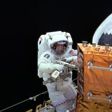 Claude Nicollier, a male European Space Agency astronaut, uses a power tool on an orange-colored storage enclosure during a spacewalk.