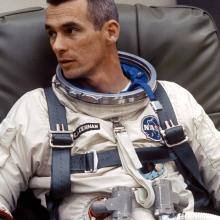 Astronaut Eugene Cernan, a white male, sits while wearing his astronaut suit without the helmet. Multiple life support tubes are seen attached to the suit.