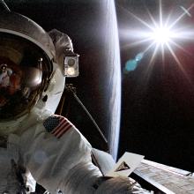 Astronaut Joseph R. Tanner, a white male astronaut, poses during a spacewalk. A silhouette of the Earth can be seen behind him.