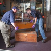 Two Museum staff members work to move a set of wooden boxes out of an exhibit.