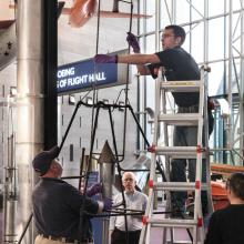 Staff members work to disassemble a primitive rocket from display in the Museum. One person is on a ladder while an other person holds the frame structure together.