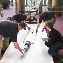 Four Museum staff members work to prepare a primitive rocket for travel away from a museum exhibit.