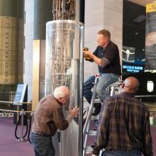 Three Museum staff members work to remove a 1940s rocket from a protective case prior to transporting the rocket.