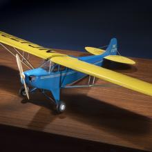 Diagonal front view of blue and yellow monoplane model with one engine represented by a wooden set of two blades.