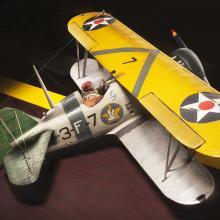 Top sidal view of yellow and silver-colored biplane model aircraft. One white star inside a blue circle is featured on each side of the top wing and identification number "3-F-7" is visible in black paint on the side of the fuselage.